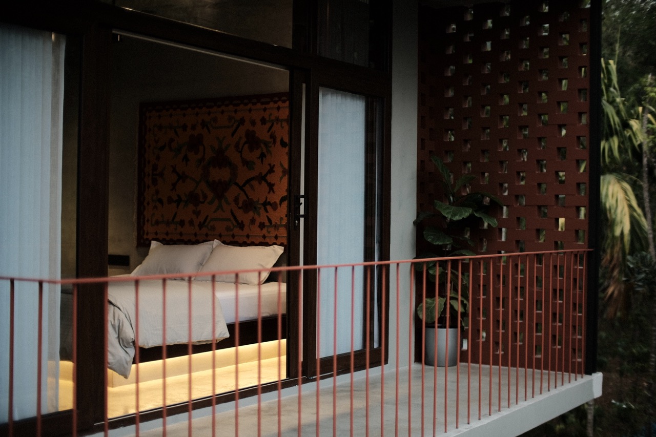Warm light spilling from a bedroom onto its balcony at dusk, beside a terracotta jali screen