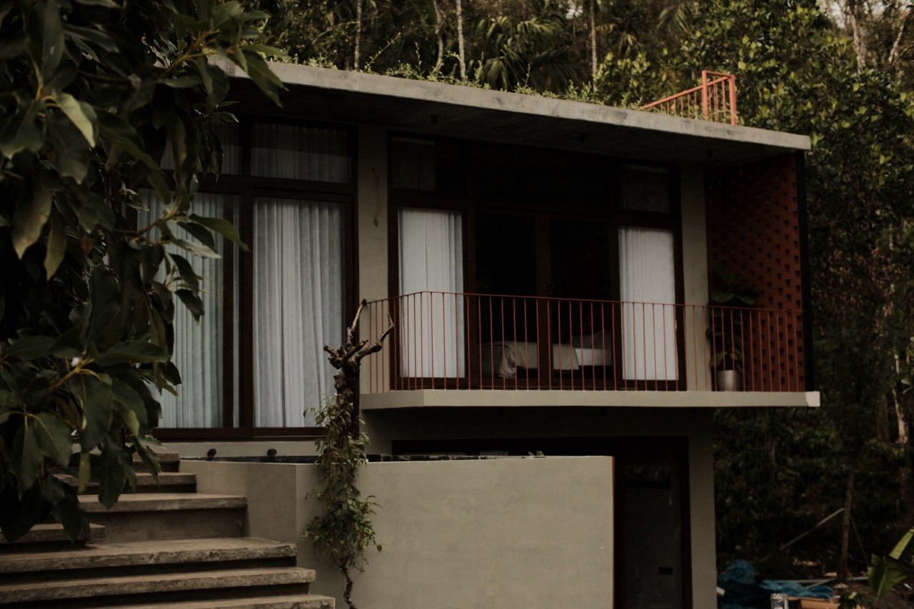 The house at dusk, upper bedroom lit behind its rust-red balcony railing