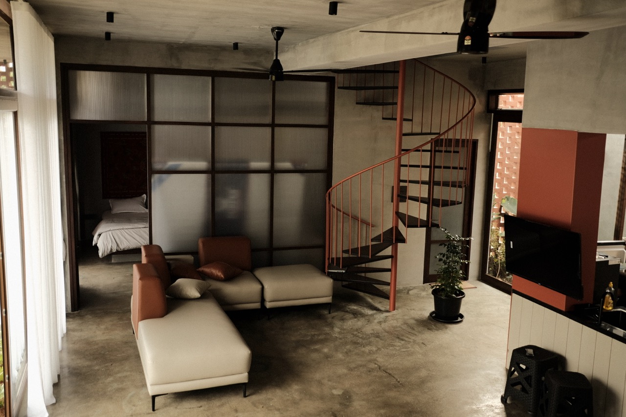 The living area with low sofa and rust-red spiral stair, opening to a bedroom beyond